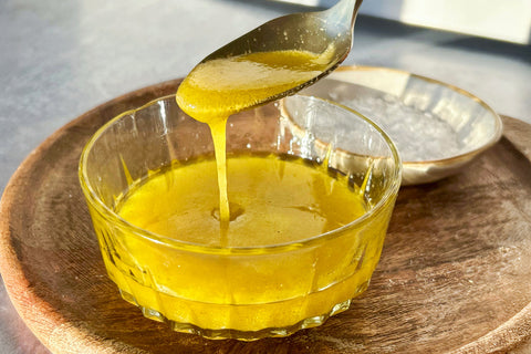 Glass bowl with yellow liquid being poured from a spoon on a wooden surface.