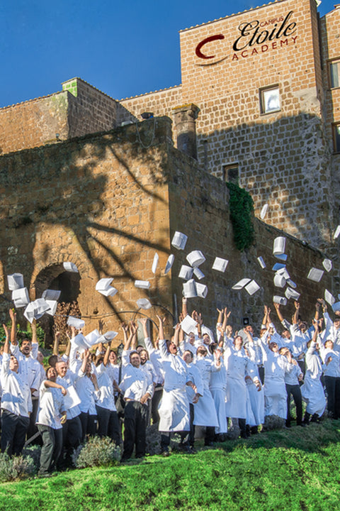 Group of people in white chef uniforms throwing hats in the air in front of a stone building with 'Cordon Bleu Etoile Academy' sign.